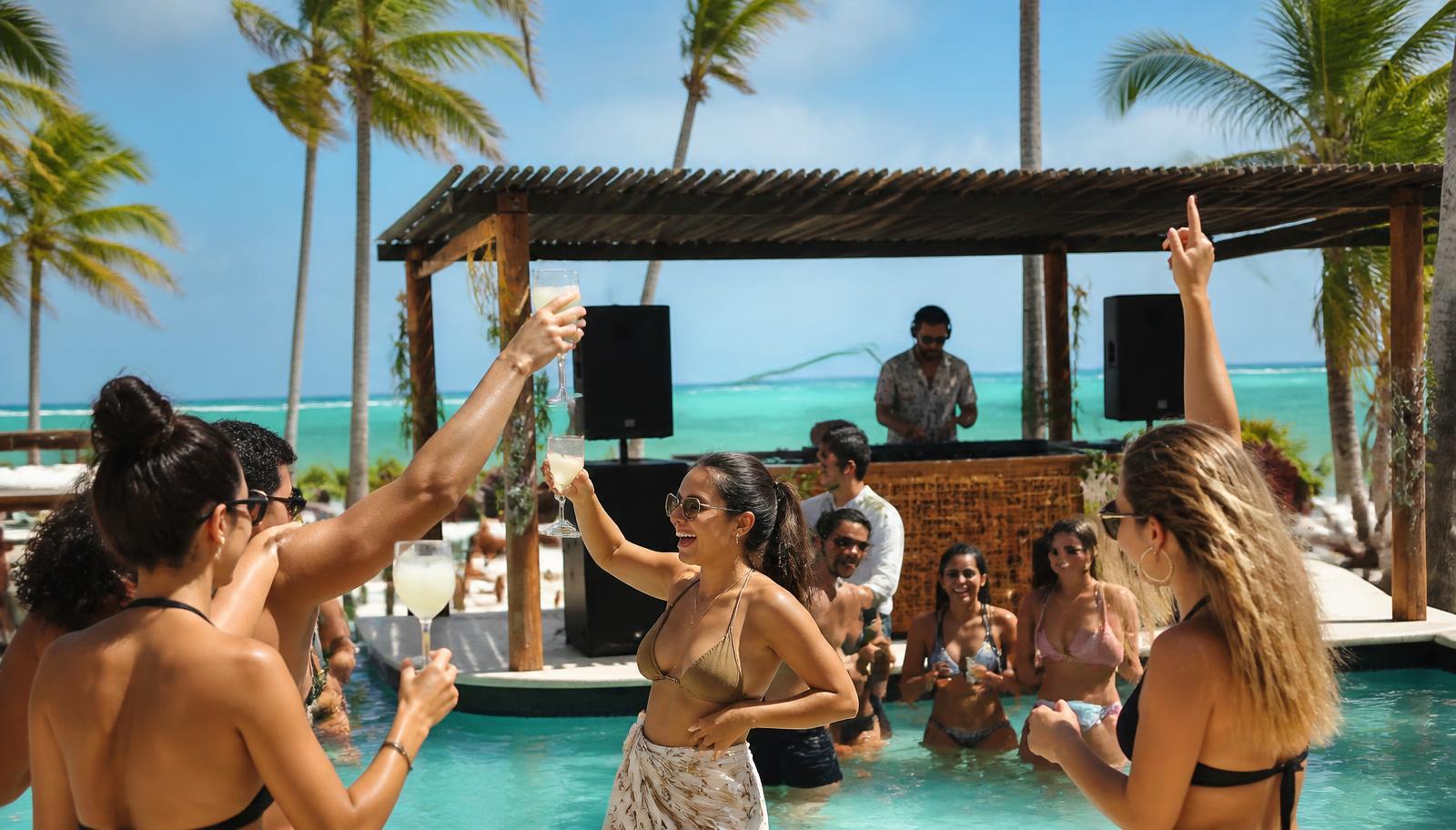 Daytime beach club pool party at an all-inclusive Punta Cana resort, friends toasting with drinks, a DJ under a palm-shaded cabana, turquoise ocean behind