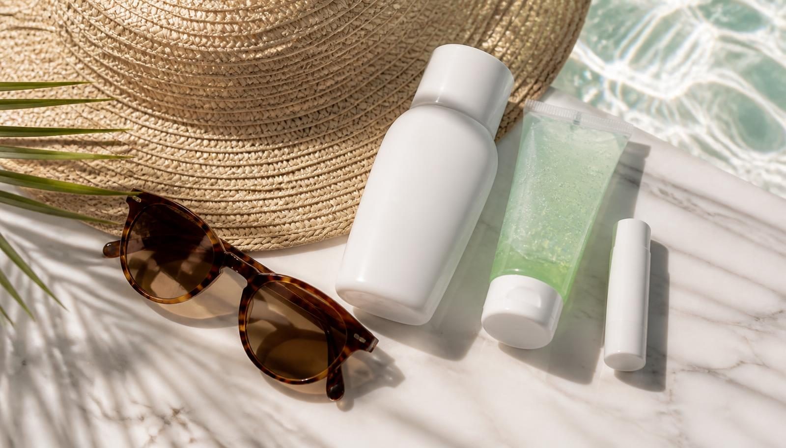 Reef-safe sunscreen bottle, aloe after-sun gel, SPF lip balm and polarized sunglasses beside a woven sun hat on white marble